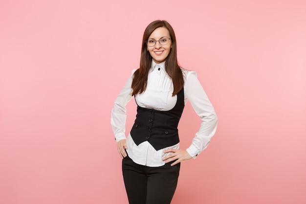 Joven alegre mujer de negocios exitosa en traje negro, camisa blanca y gafas que se encuentran aisladas sobre fondo rosa pastel. Jefa. Concepto de riqueza de carrera de logro. Copie el espacio para publicidad.