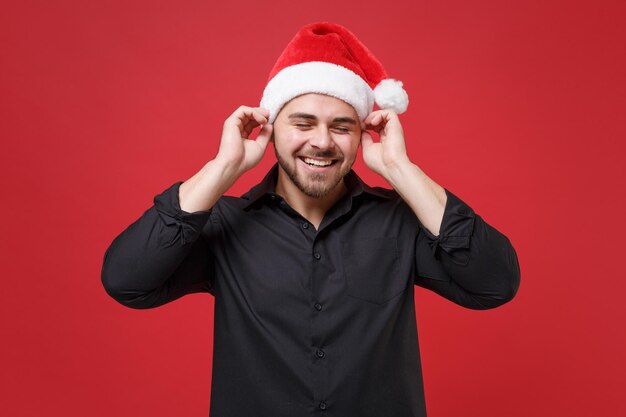 Un joven y alegre Papá Noel barbudo con camisa negra clásica, sombrero de Navidad aislado de fondo rojo. Concepto de vacaciones de celebración de año nuevo feliz. Simulacros de espacio de copia. Poner las manos en la cabeza, manteniendo los ojos cerrados.