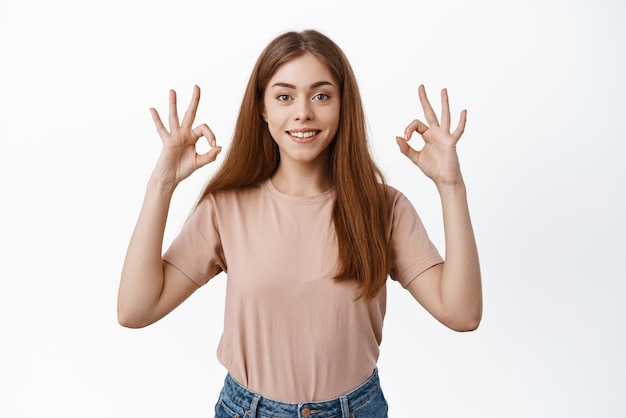 Una joven alegre que sonríe asiente con aprobación y muestra señales de estar bien, da permiso, deja comentarios positivos sobre el producto, dice sí o está bien, de pie contra el fondo blanco