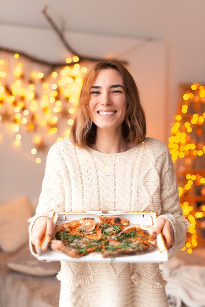 La joven alegre que sonríe a la cámara vertical sostiene una caja de pizza.