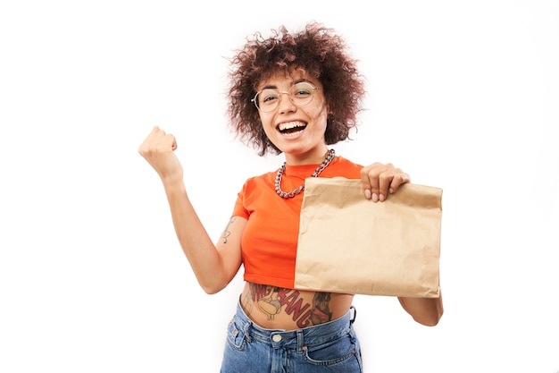 Joven alegre con rizos afro en ropa naranja con paquete de artesanía aislado sobre fondo blanco de estudio, entrega de alimentos, bolsa ecológica, chica caucásica kazaja con regalo