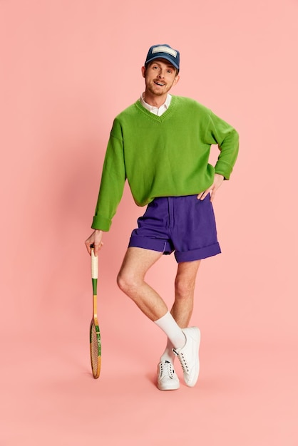 Un joven alegre con ropa deportiva vintage posando apoyado en una raqueta de tenis aislado sobre un fondo rosa Club deportivo