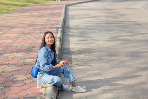 Una joven alegre sentada en el parque de la ciudad sostiene un teléfono móvil con una botella de agua colocada en el concepto de estilo de vida lateral