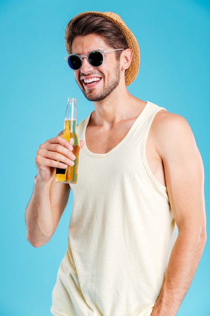 Joven alegre con sombrero y gafas de sol de pie y bebiendo cerveza sobre la pared azul