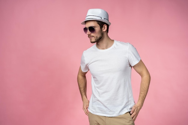 Joven alegre con sombrero y gafas de sol de pie y sonriendo sobre fondo rosa.