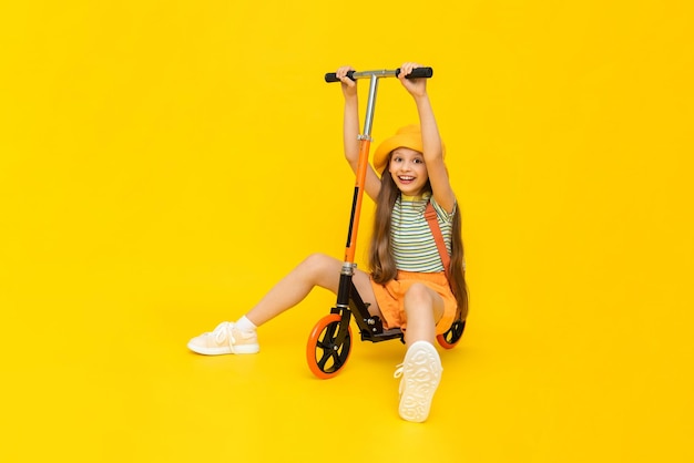 Una joven alegre con sombrero y pantalones cortos está jugando en un scooter de dos ruedas Recreación activa de la ciudad para niños Fondo aislado amarillo