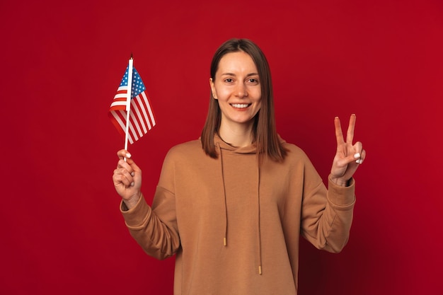 Una joven alegre sostiene la bandera de Estados Unidos y un gesto de paz con dos dedos