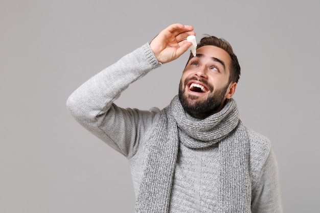 Joven alegre con suéter gris, bufanda aislada en un retrato de estudio de fondo de pared gris. Estilo de vida saludable, tratamiento de enfermedades enfermas, concepto de temporada fría. Simulacros de espacio de copia. Mantenga las gotas para los ojos.