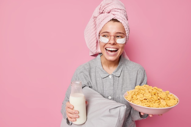 Una joven alegre tiene un desayuno saludable, sostiene un plato con cereales y una botella de leche, se ríe felizmente vestida con ropa de dormir, se somete a procedimientos de belleza después de dormir, posa contra una pared rosa.