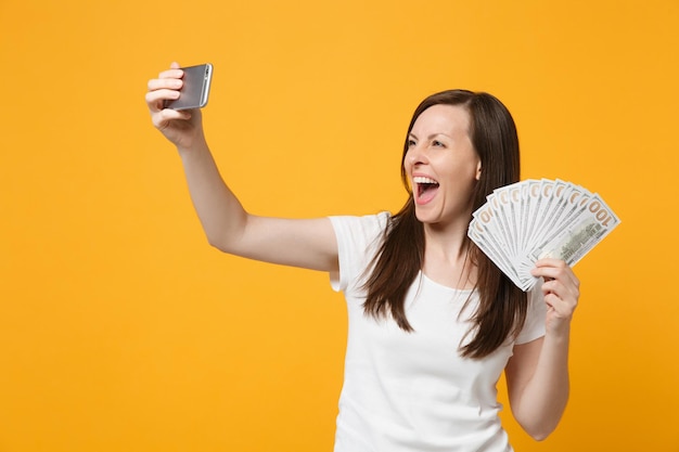 Una joven alegre vestida de blanco haciendo selfie en un teléfono móvil, sosteniendo un abanico de dinero en efectivo en billetes en dólares aislados en un fondo naranja amarillo. Concepto de estilo de vida de las personas. Simulacros de espacio de copia.