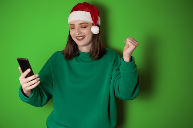 Una joven alegre vestida con un suéter verde y un sombrero de santa sosteniendo un teléfono móvil en la mano haciendo un gesto de ganador aislado en un fondo verde Concepto de vacaciones de feliz año nuevo 2023