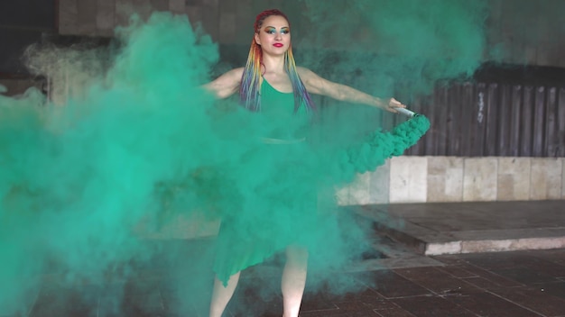 Una joven alegre con un vestido de primavera verde brillante con maquillaje con lentejuelas y trenzas africanas de arco iris. Ella se para cerca de un edificio y lanza humo verde en la calle.