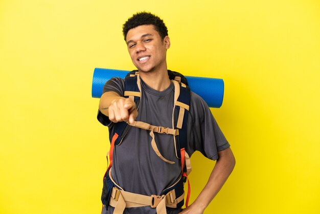 Joven alpinista afroamericano con una gran mochila aislada sobre fondo amarillo apuntando hacia el frente con expresión feliz