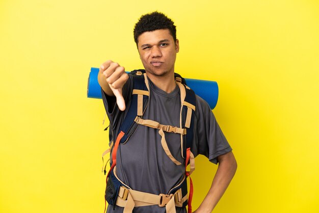 Joven alpinista afroamericano con una gran mochila aislada sobre fondo amarillo que muestra el pulgar hacia abajo con expresión negativa