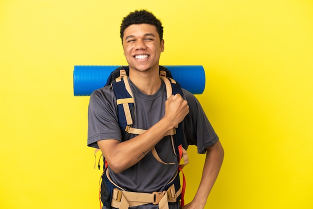 Joven alpinista afroamericano con una gran mochila aislado sobre fondo amarillo celebrando una victoria
