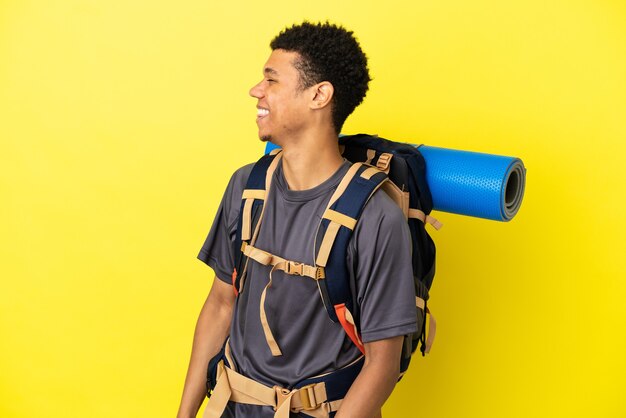 Joven alpinista afroamericano con una gran mochila aislado sobre fondo amarillo riendo en posición lateral