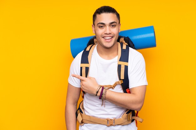 Joven alpinista asiático con una mochila grande aislada en amarillo dedo acusador hacia el lado