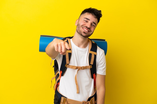 Joven alpinista hombre caucásico con una gran mochila aislada sobre fondo amarillo apuntando al frente con expresión feliz