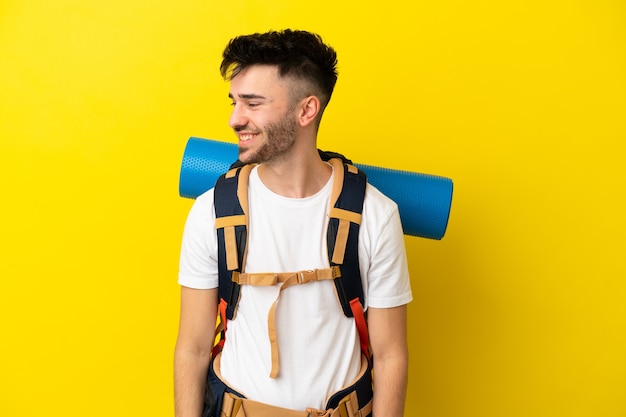 Joven alpinista hombre caucásico con una gran mochila aislada sobre fondo amarillo mirando hacia el lado y sonriendo