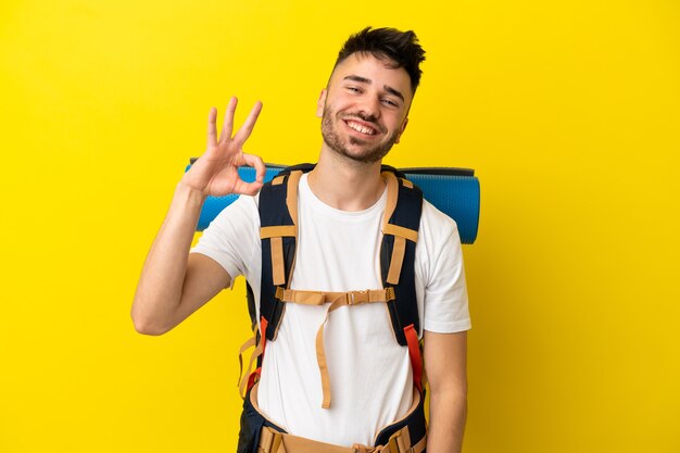 Joven alpinista hombre caucásico con una gran mochila aislada sobre fondo amarillo mostrando signo ok con los dedos