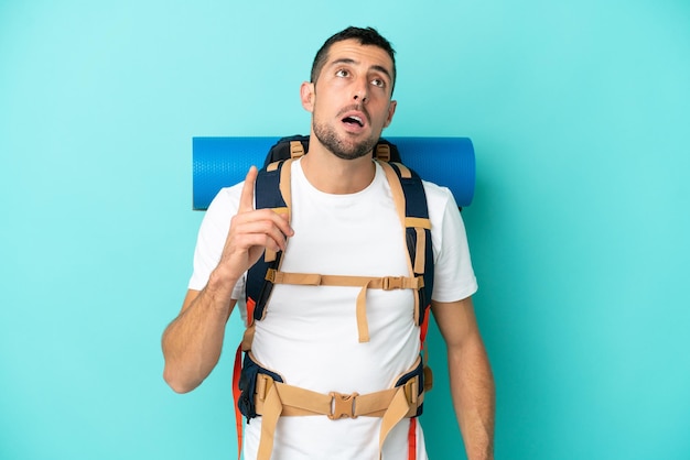 Joven alpinista hombre caucásico con una gran mochila aislada sobre fondo azul pensando en una idea apuntando con el dedo hacia arriba