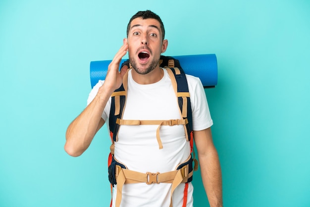 Joven alpinista hombre caucásico con una gran mochila aislada sobre fondo azul con sorpresa y expresión facial conmocionada