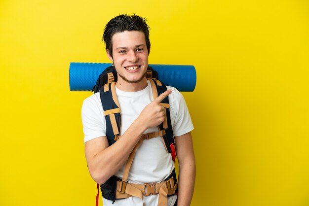 Joven alpinista ruso con una gran mochila aislado sobre fondo amarillo apuntando hacia el lado para presentar un producto