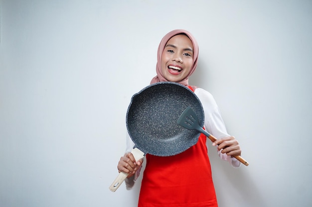 Joven ama de casa asiática con delantal de cocina cocinando y sosteniendo una sartén y una espátula