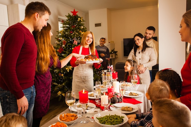 Joven ama de casa atractiva sosteniendo un plato con pavo asado recién cocinado mostrando su plato favorito a un grupo de amigos en la cena de Navidad