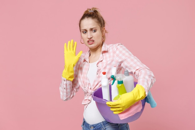 Una joven ama de casa disgustada con guantes de goma sostiene un lavabo con botellas de detergente lavando limpiadores haciendo tareas domésticas aisladas en un fondo rosado. Concepto de limpieza. Mostrando gesto de parada con la palma.