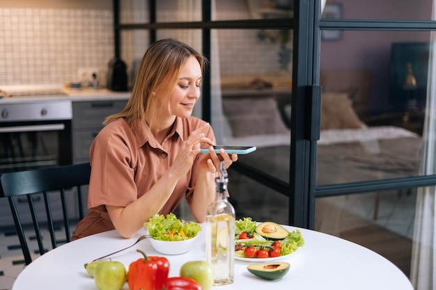 Una joven ama de casa feliz y despreocupada usa un smartphone para tomar fotos de verduras en la mesa de la cocina. Mujer tomando fotos de comida vegetariana en un teléfono celular inteligente.