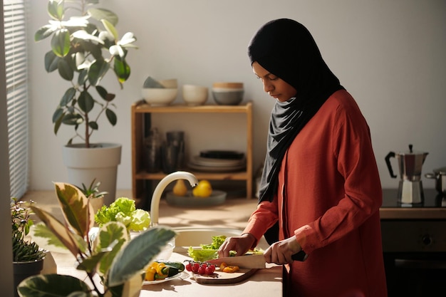 Joven ama de casa musulmana con hijab y ropa casual cortando verduras frescas a bordo para ensalada mientras está de pie en el mostrador de la cocina