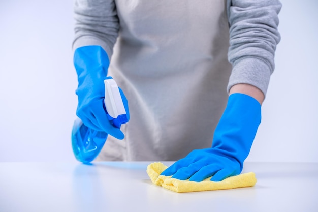 Joven ama de llaves está limpiando la mesa blanca en el delantal con guantes azules, limpiador en aerosol, trapo amarillo mojado, primer plano, espacio de copia, concepto de diseño en blanco