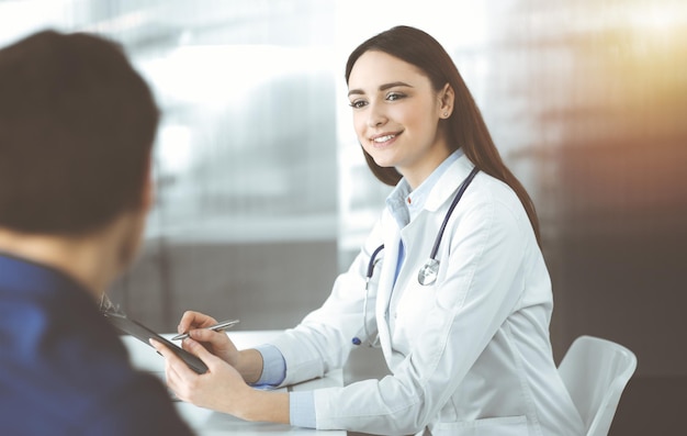 Una joven y amable doctora está escuchando a su paciente, mientras se sientan juntas en una clínica soleada. Conceptos de medicina y salud.