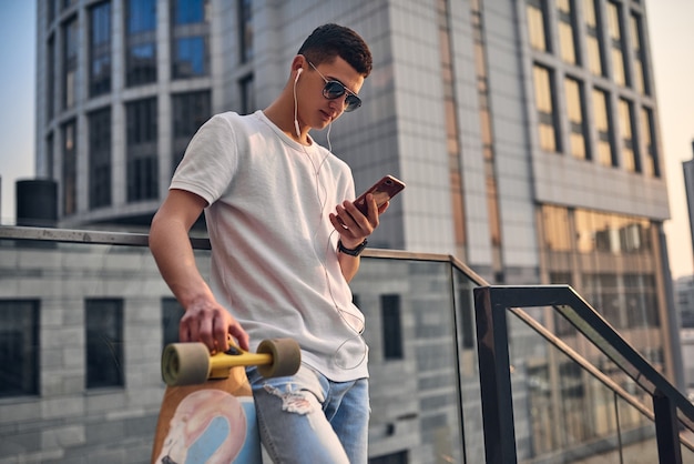 Joven americano caucásico con gafas en los escalones con un longboard contra el fondo