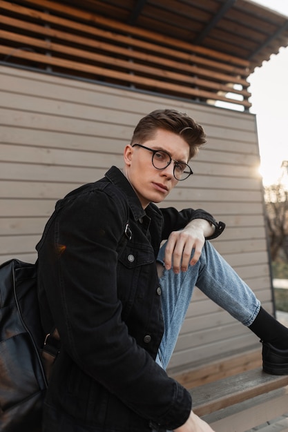 Joven americano en ropa de mezclilla casual de moda en gafas elegantes con mochila negra de moda