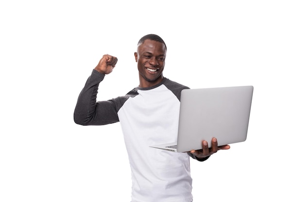 Un joven americano vestido con un suéter blanco y negro trabaja felizmente en él hombre sosteniendo una computadora portátil