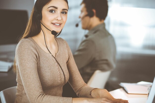 Una joven amigable con auriculares está hablando con el cliente de una empresa, mientras está sentada en el escritorio de una oficina moderna junto con su colega. Operadores de call center en el trabajo.