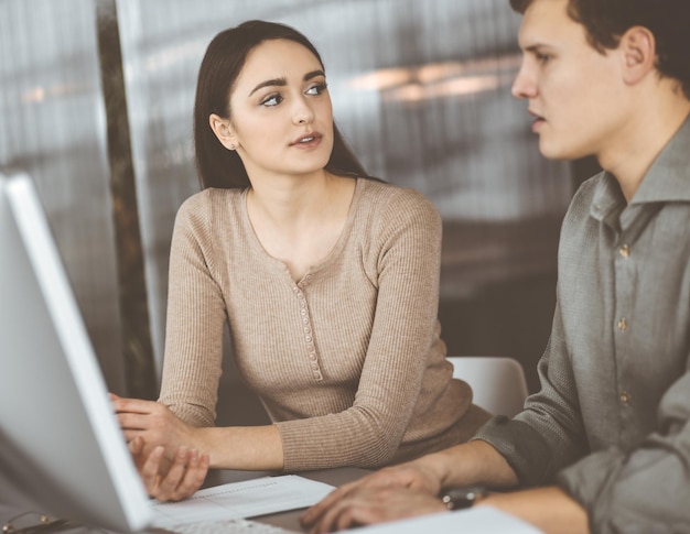 Un joven y amigable hombre de negocios y programador con una camisa verde está trabajando en una computadora, mientras se sientan juntos en el escritorio con una colega en una oficina moderna. Centrarse en la mujer. Concepto de éxito