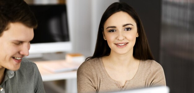 Un joven y amigable hombre de negocios y programador con una camisa verde está trabajando en una computadora, mientras se sientan juntos en el escritorio con una colega en una oficina moderna. Centrarse en la mujer. Concepto de éxito