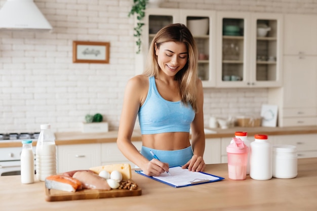 Joven amigable sonriendo y escribiendo en su registro de nutrición mientras está de pie cerca de la mesa con productos alimenticios