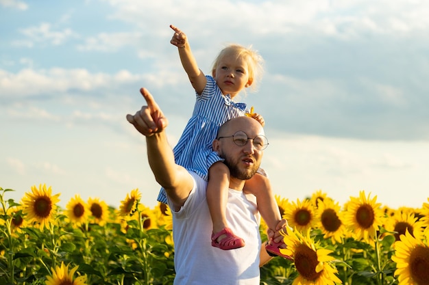 Joven con anteojos sostiene a un niño sobre sus hombros y señala con la mano algo en un campo de girasoles