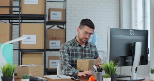 Un joven con anteojos usa una computadora de escritorio una mujer clasifica artículos en cajas de cartón Los empleados preparan pedidos de venta en la tienda en línea