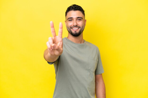 Joven apuesto árabe aislado de fondo amarillo sonriendo y mostrando el signo de la victoria