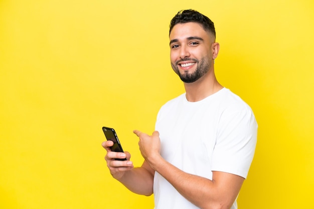 Joven apuesto árabe aislado de fondo amarillo usando teléfono móvil y apuntando hacia atrás