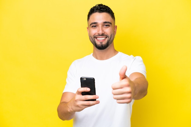 Joven apuesto árabe aislado de fondo amarillo usando teléfono móvil mientras hace pulgares hacia arriba