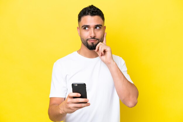 Joven apuesto árabe aislado de fondo amarillo usando teléfono móvil y pensando