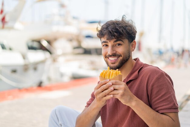 Foto joven apuesto árabe al aire libre tomando patatas fritas con expresión feliz