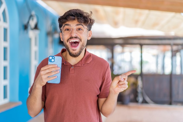 Joven apuesto árabe usando un teléfono móvil al aire libre sorprendido y señalando con el dedo hacia un lado