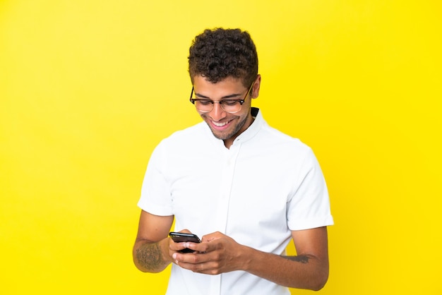 Joven apuesto brasileño aislado de fondo amarillo enviando un mensaje con el móvil